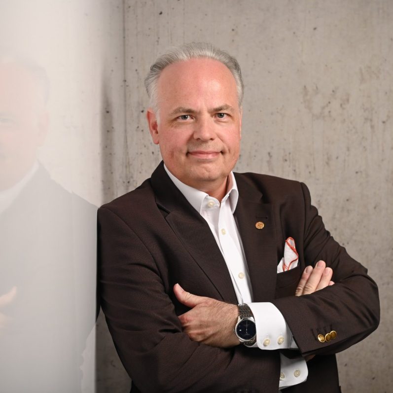 Mag.phil. Arne Behr Männlicher Psychotherapeut mit verschränkten Armen, grauem Haar und braunem Sakko vor Betonwand.