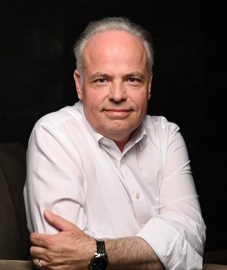 Mag.phil. Arne Behr Männlicher Psychotherapeut mit verschränkten Armen, grauem Haar und braunem Sakko vor Betonwand.